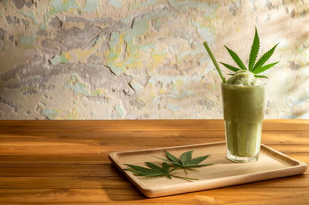 A glass of cannabis smoothie with cannabis leaves on a wooden tray on a wooden table